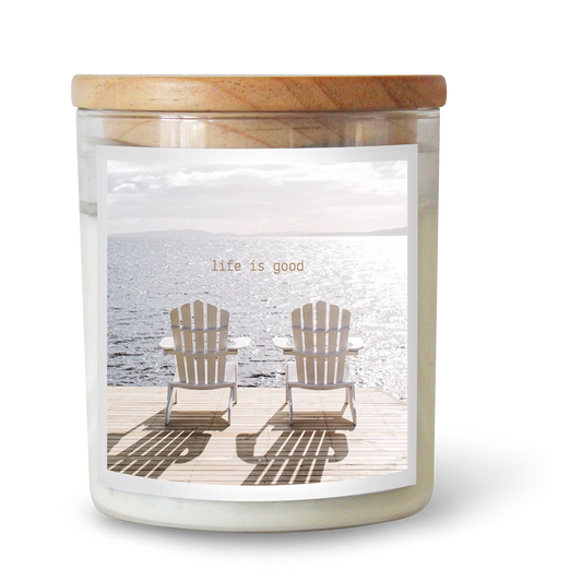 The Commonfolk scented white candle features a label with two wooden chairs on a beach, along with the text 'Life is good' above the image.
