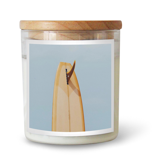 The Commonfolk scented candle comes in a white soy wax container, with a wooden lid and a label featuring a minimalist illustration of a surfboard.