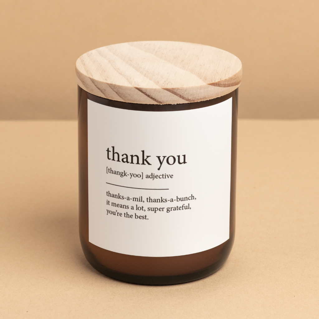 The Commonfolk scented candle has a white label that reads 'thank you' in different fonts, topped with a wooden lid on a beige background.