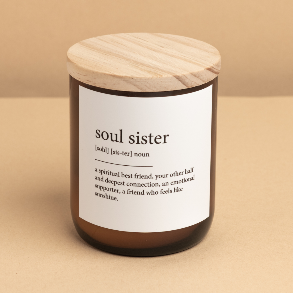 The Commonfolk scented candle, with an amber jar and a wooden lid, labeled 'soul sister' with a definition next to it, is placed on a beige background.