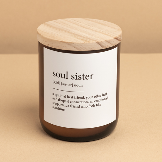 The Commonfolk scented candle, with an amber jar and a wooden lid, labeled 'soul sister' with a definition next to it, is placed on a beige background.