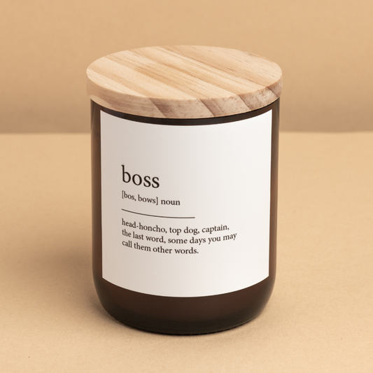 The Commonfolk scented candle has a white label that defines the word 'boss'. The candle is in a dark container with a wooden lid.