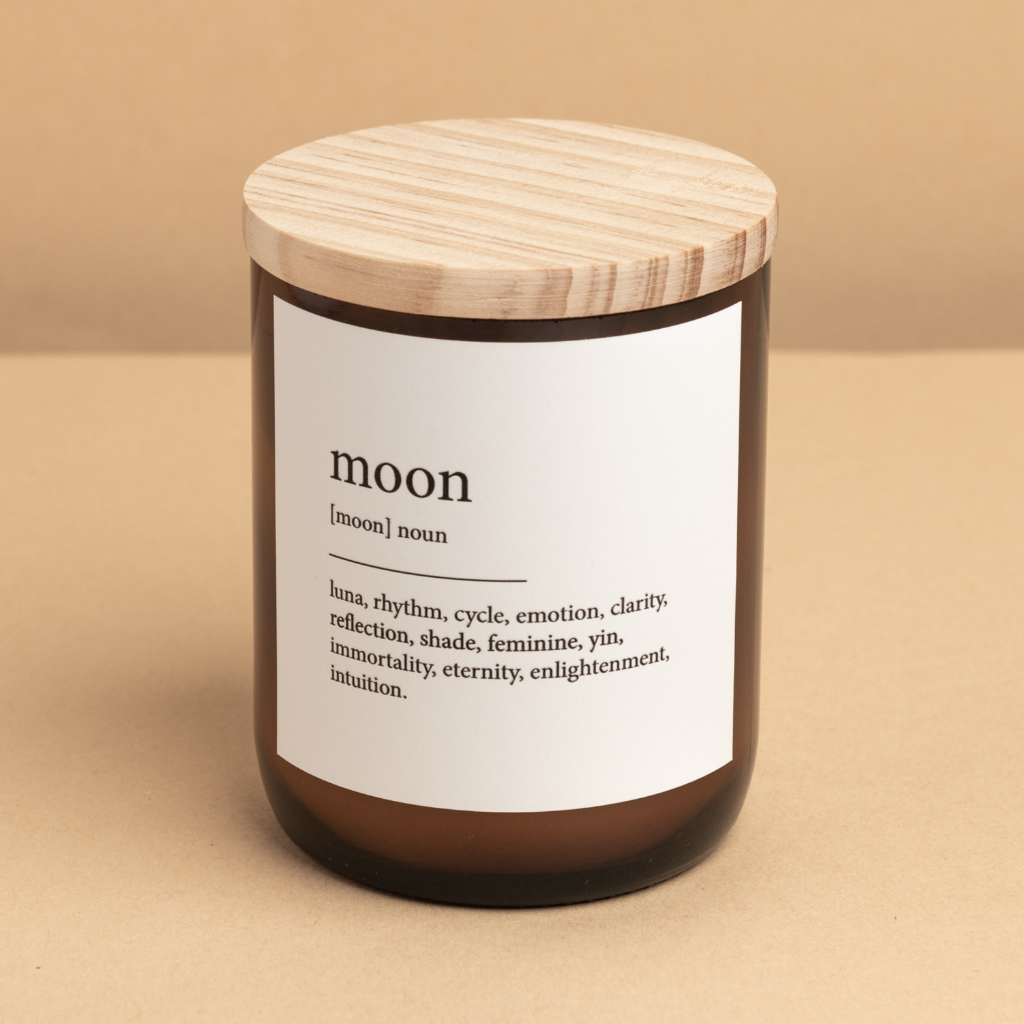 The Commonfolk scented amber glass candle jar with a wooden lid, labeled 'moon' with a definition in brackets, is set against a beige background.