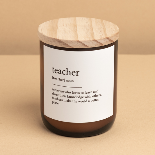 The Commonfolk scented soy wax candle in an amber jar with a wooden lid, labeled 'teacher,' is defined as 'someone who loves to learn and share their knowledge with others.'