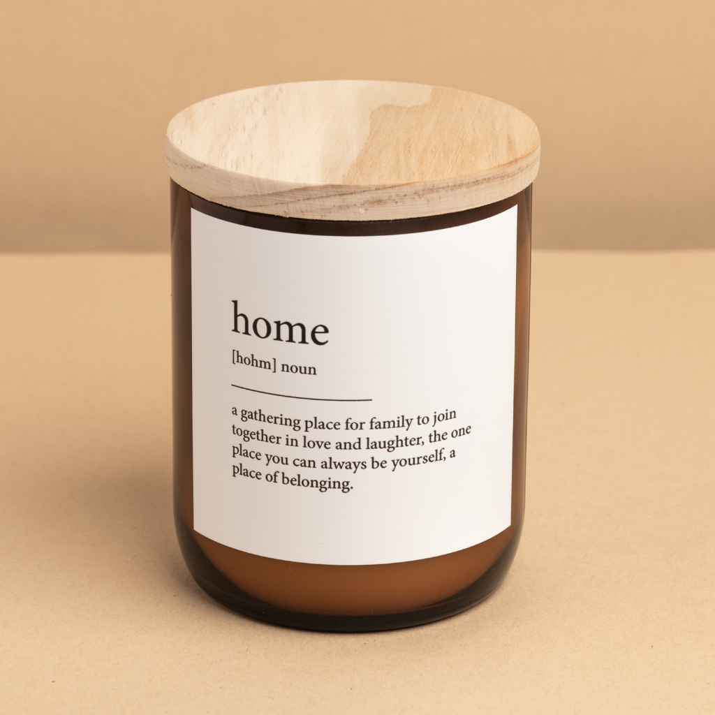 The Commonfolk scented candle, with an amber glass jar and a wooden lid, is labeled 'home' with a definition in brackets, placed on a beige background.