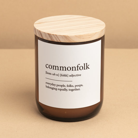 The Commonfolk scented medium-sized amber jar candle with a wooden lid, labeled 'commonfolk' with a definition of 'everyday people, folk, populace' on the label.