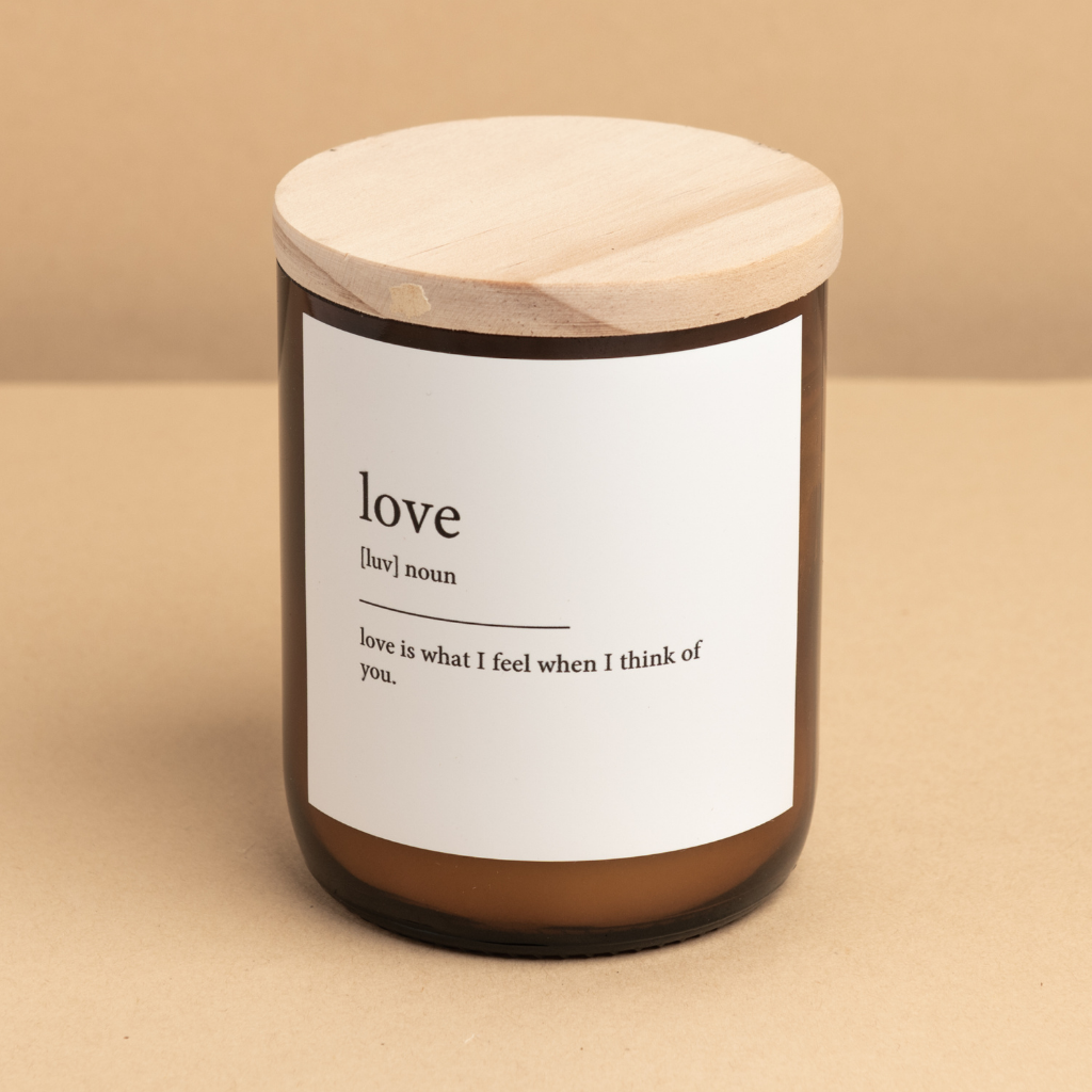The Commonfolk scented candle has a wooden lid and a label that defines 'love,' written in brackets above the word 'love.' The candle is placed against a beige background.