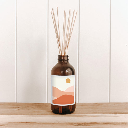 The Commonfolk room diffuser with natural bamboo reed sticks in an amber glass bottle with a label featuring a sun and desert landscape.