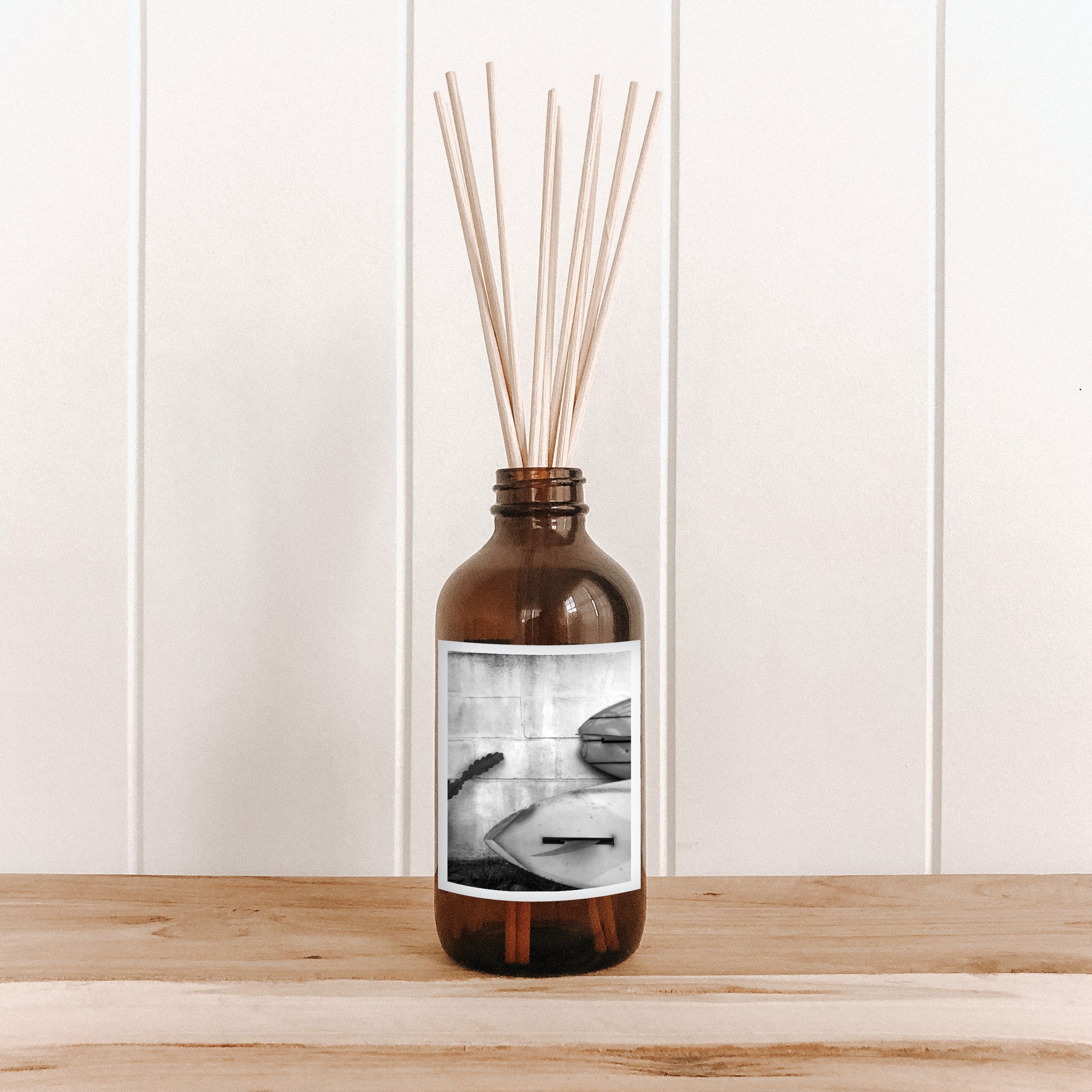 The Commonfolk room diffuser with natural bamboo reed sticks in a clear glass bottle features a black and white photo of a surfboard on the label.