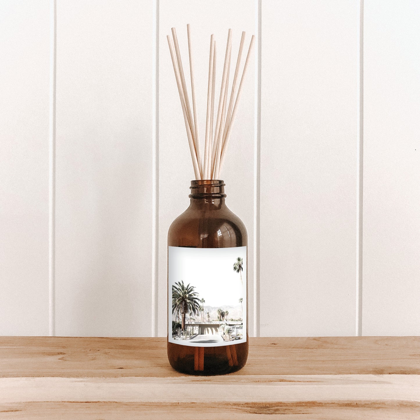 The Commonfolk room diffuser with natural bamboo reed sticks in a brown bottle with a white label featuring a palm tree and house illustration.