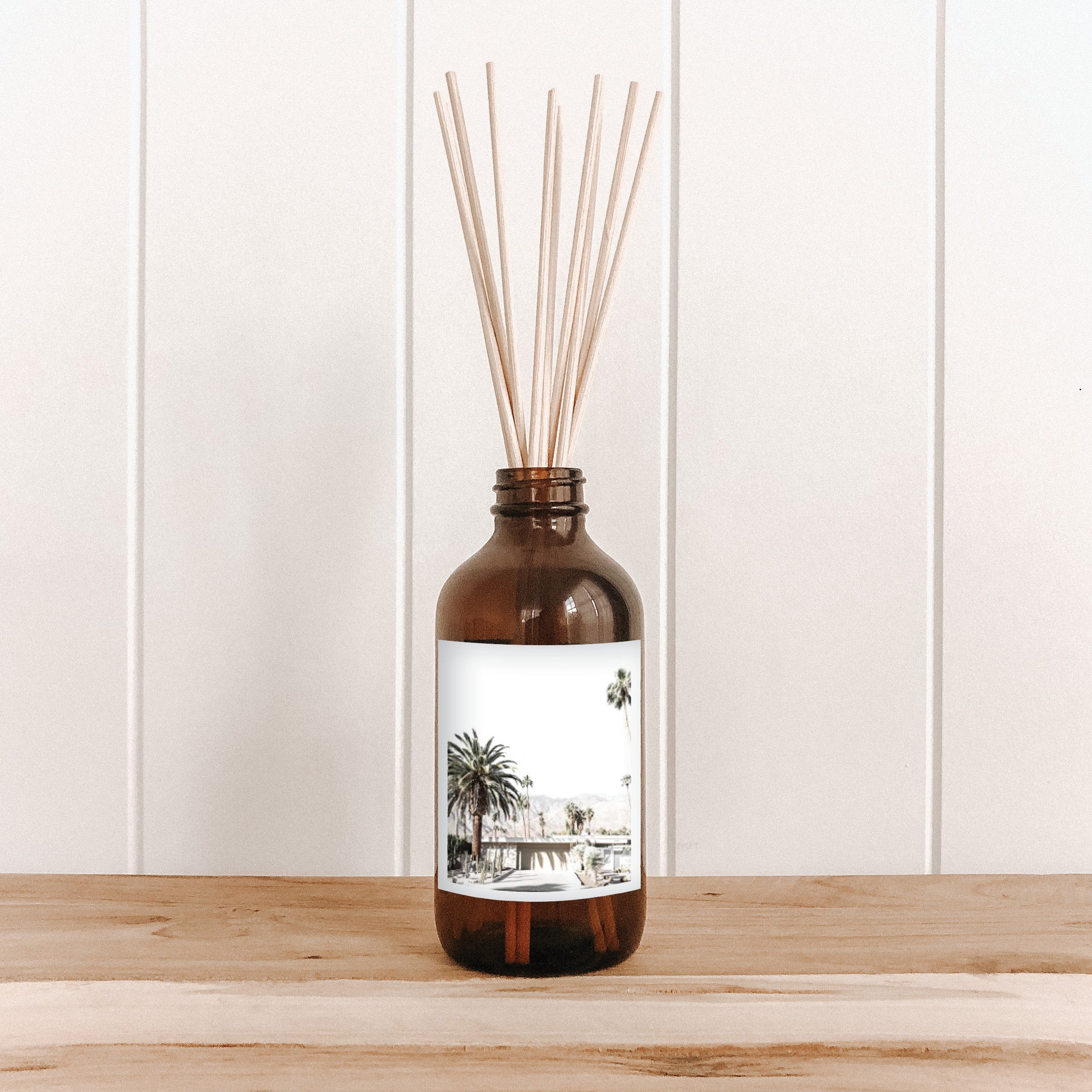The Commonfolk room diffuser with natural bamboo reed sticks in a brown bottle with a white label featuring a palm tree and house illustration.