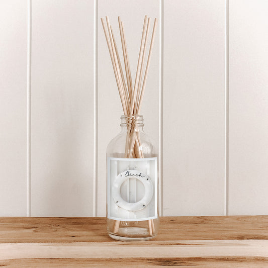 The Commonfolk clear glass room diffuser bottle with natural bamboo reed sticks inserted, set on a wooden surface against a white background, labeled 'Beach'.
