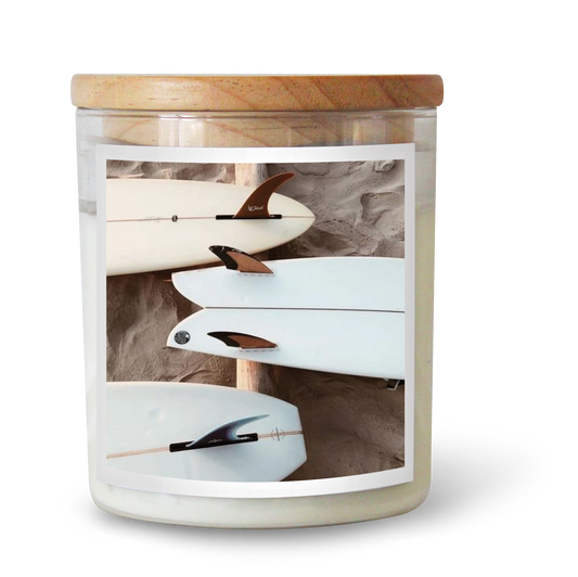 The Commonfolk scented candle has a white label inside a clear jar, featuring a wooden lid. The label shows a white surfboard.