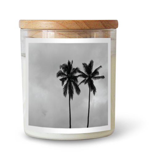 The Commonfolk scented candle features a label with two palm trees against a grey background and has a wooden lid on top.