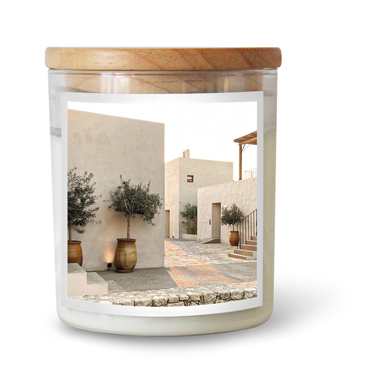 The Commonfolk scented candle features white wax, a clear glass jar, and a wooden lid, with an image of a serene street scene that includes buildings and a tree.
