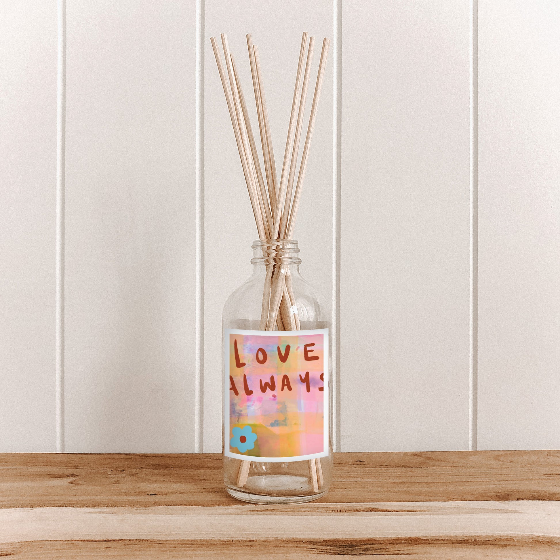 The Commonfolk room diffuser bottle with 'LOVE ALWAYS' text design, containing natural bamboo reed sticks, is placed on a wooden surface against a white background.