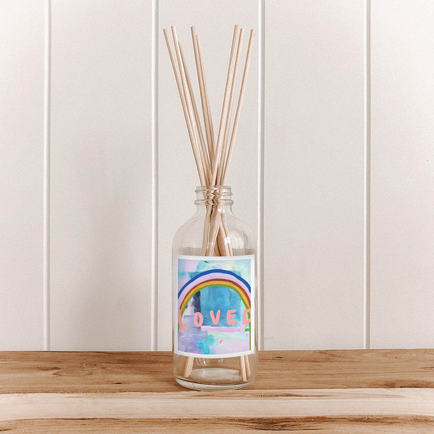 The Commonfolk room diffuser bottle, with a label featuring a rainbow and the word 'LOVED', filled with natural bamboo reed sticks, is placed on a wooden surface against a white background.