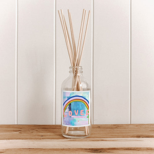 The Commonfolk room diffuser bottle, with a label featuring a rainbow and the word 'LOVED', filled with natural bamboo reed sticks, is placed on a wooden surface against a white background.