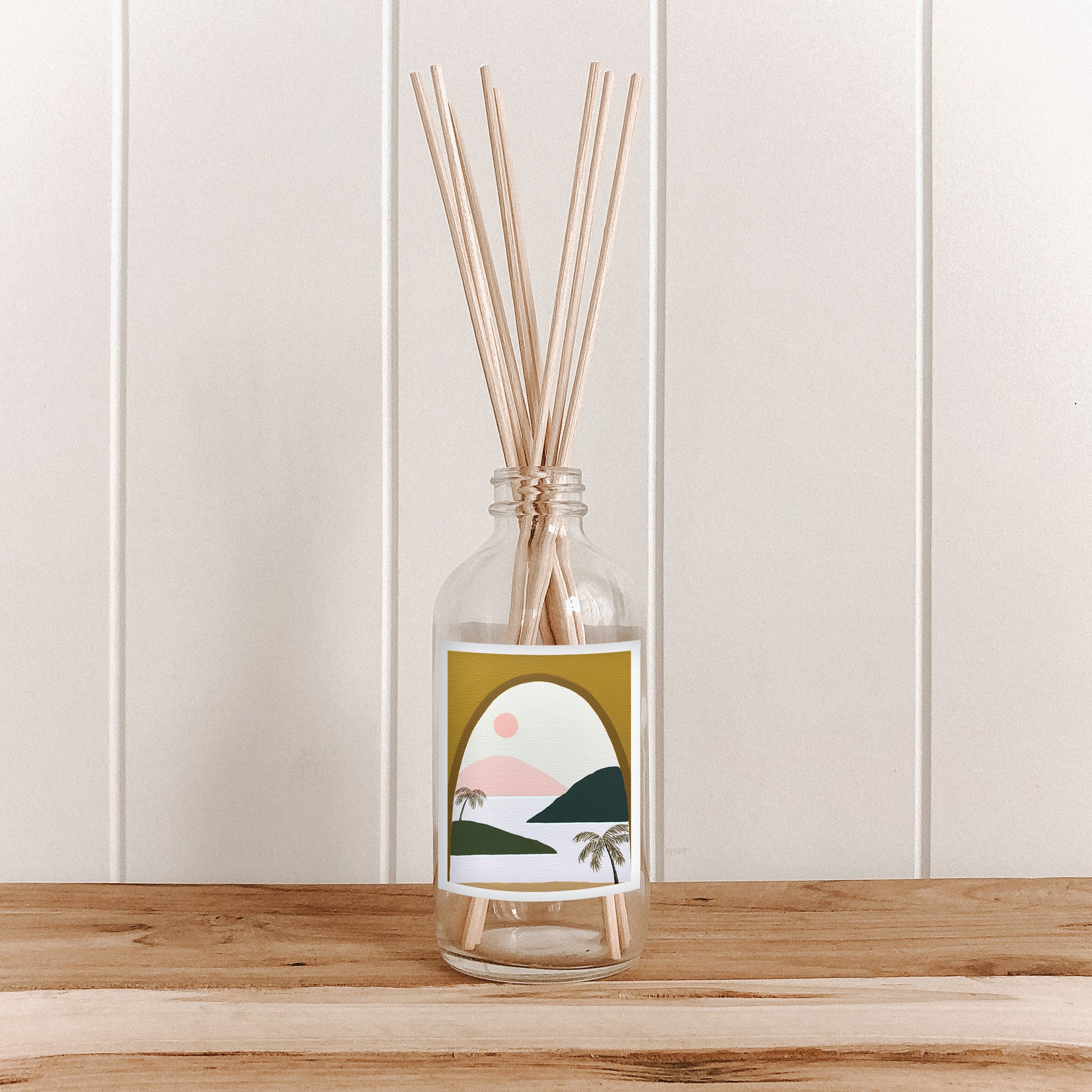 The Commonfolk room diffuser, a clear bottle with a label featuring an artistic illustration, is placed on a wooden surface with natural bamboo reed sticks inside the bottle.