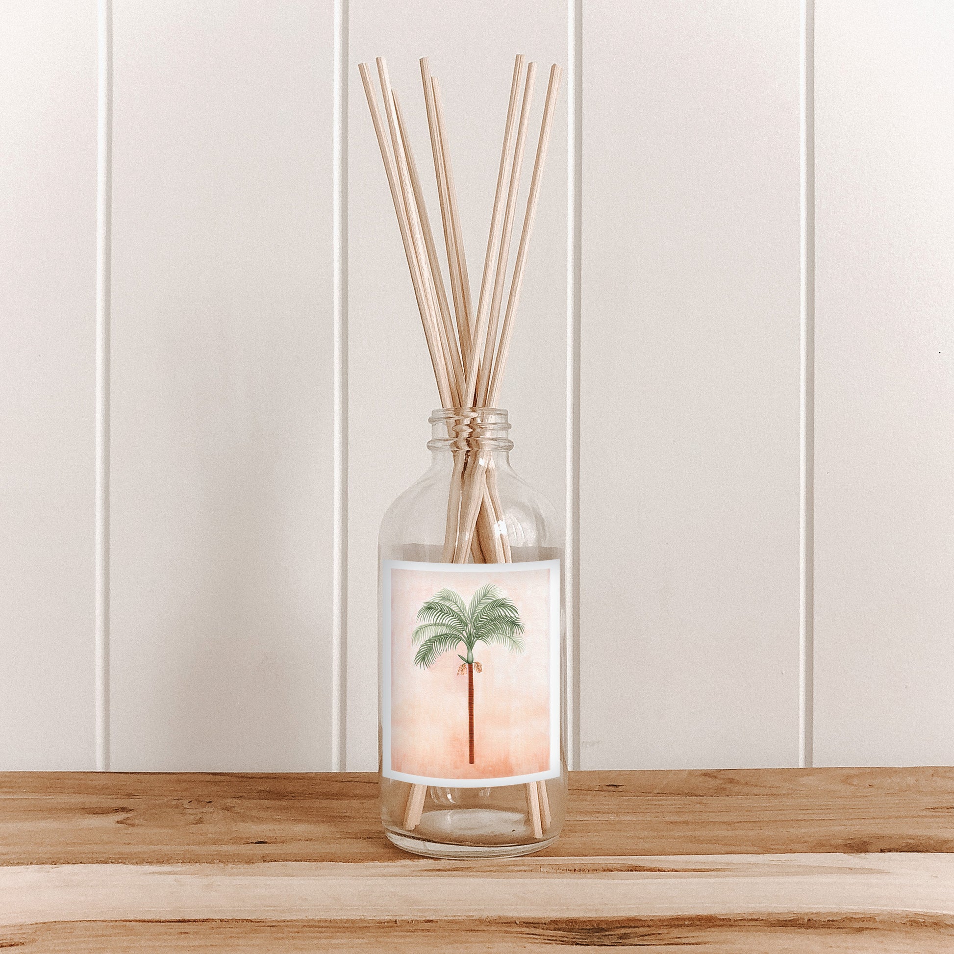 The Commonfolk room diffuser with natural bamboo reed sticks in a clear glass bottle featuring a palm design on the label is placed on a wooden surface against a white background.