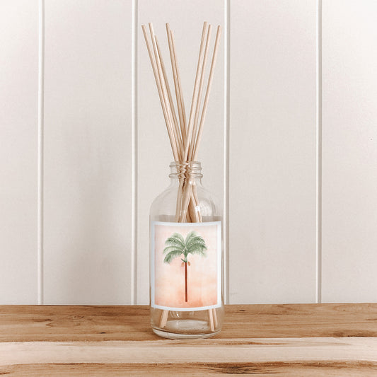 The Commonfolk room diffuser with natural bamboo reed sticks in a clear glass bottle featuring a palm design on the label is placed on a wooden surface against a white background.