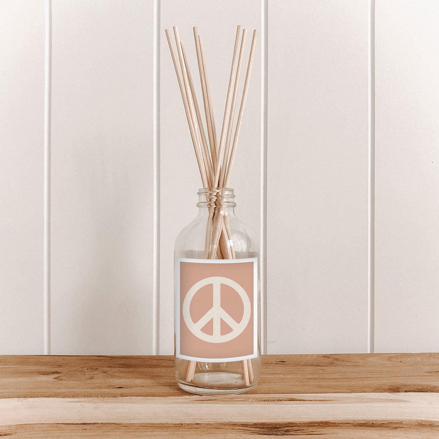 A room diffuser with natural bamboo reed sticks in a clear glass bottle, featuring a pink label with a peace sign.