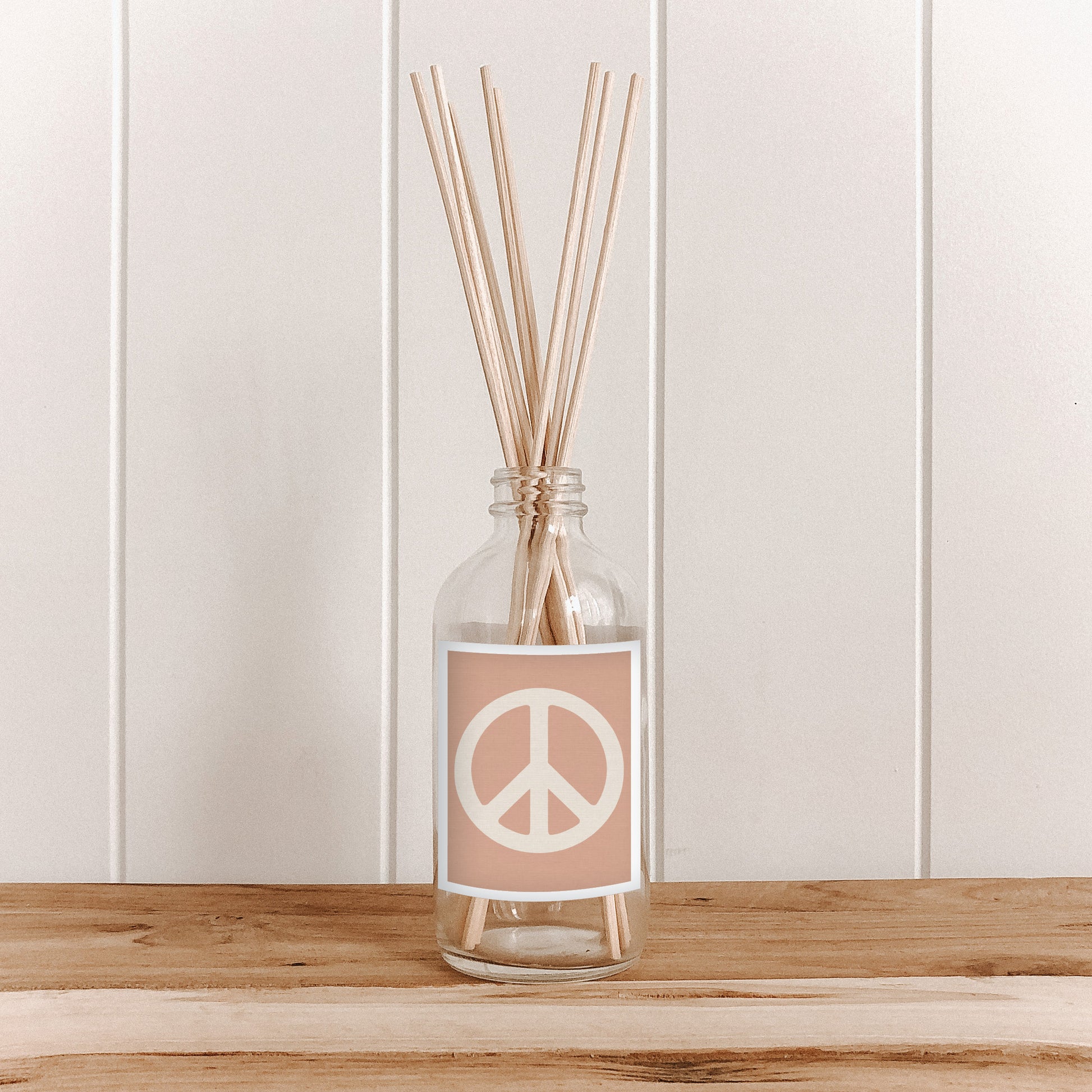 A room diffuser with natural bamboo reed sticks in a clear glass bottle, featuring a pink label with a peace sign.