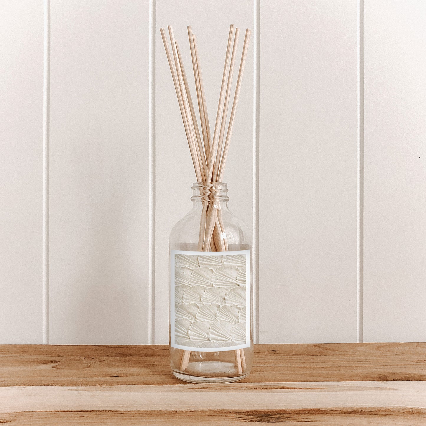 The Commonfolk clear glass room diffuser bottle with a white label, containing natural bamboo reed sticks, is placed on a wooden surface against a white background.