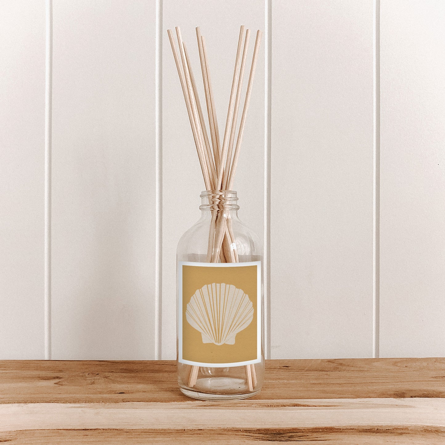 The Commonfolk room diffuser with natural bamboo reed sticks is in a clear glass bottle with a yellow label featuring a shell design. The bottle is placed on a wooden surface with a white background.