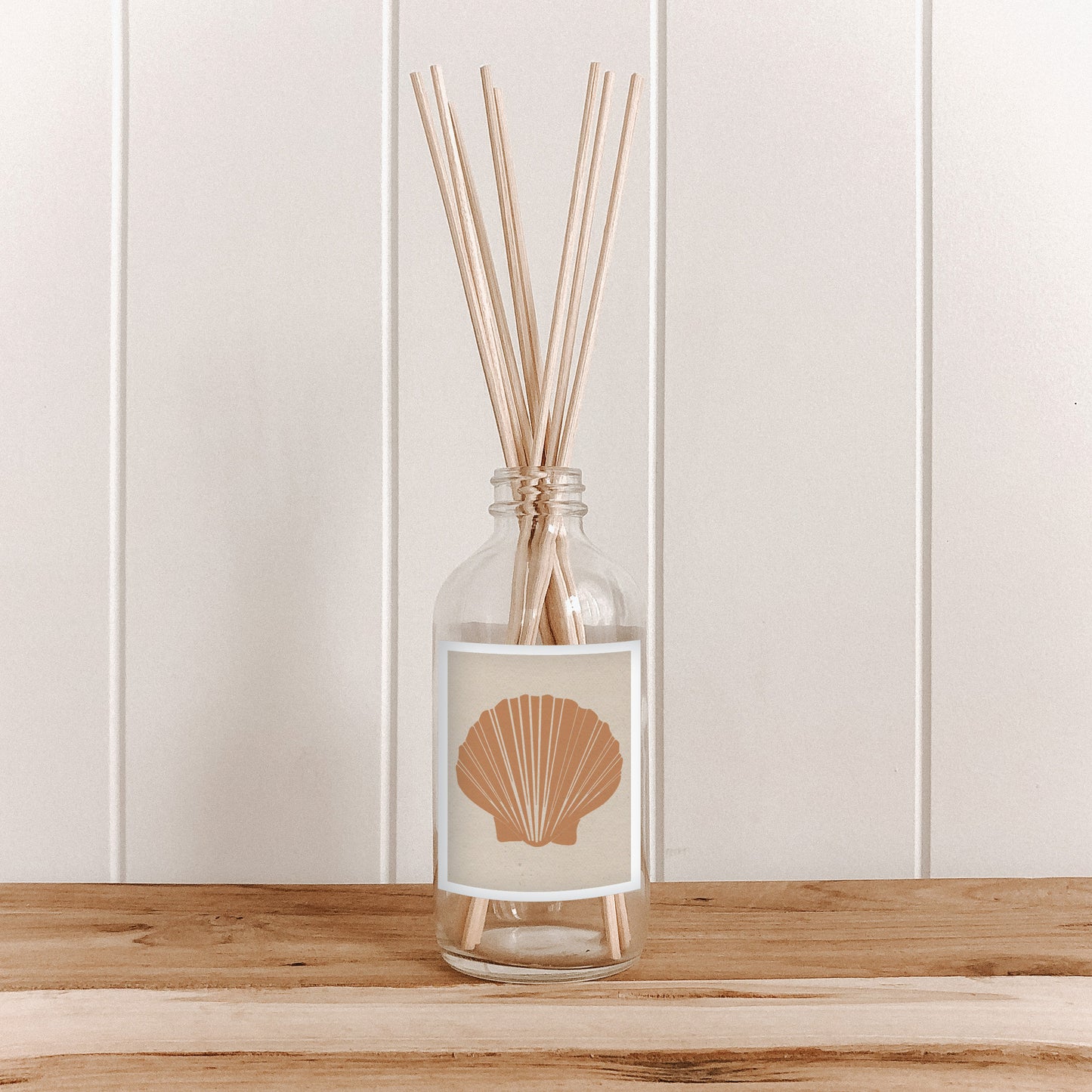 The Commonfolk room diffuser with natural bamboo reed sticks in a clear glass bottle with a tan label featuring a fan shell design.