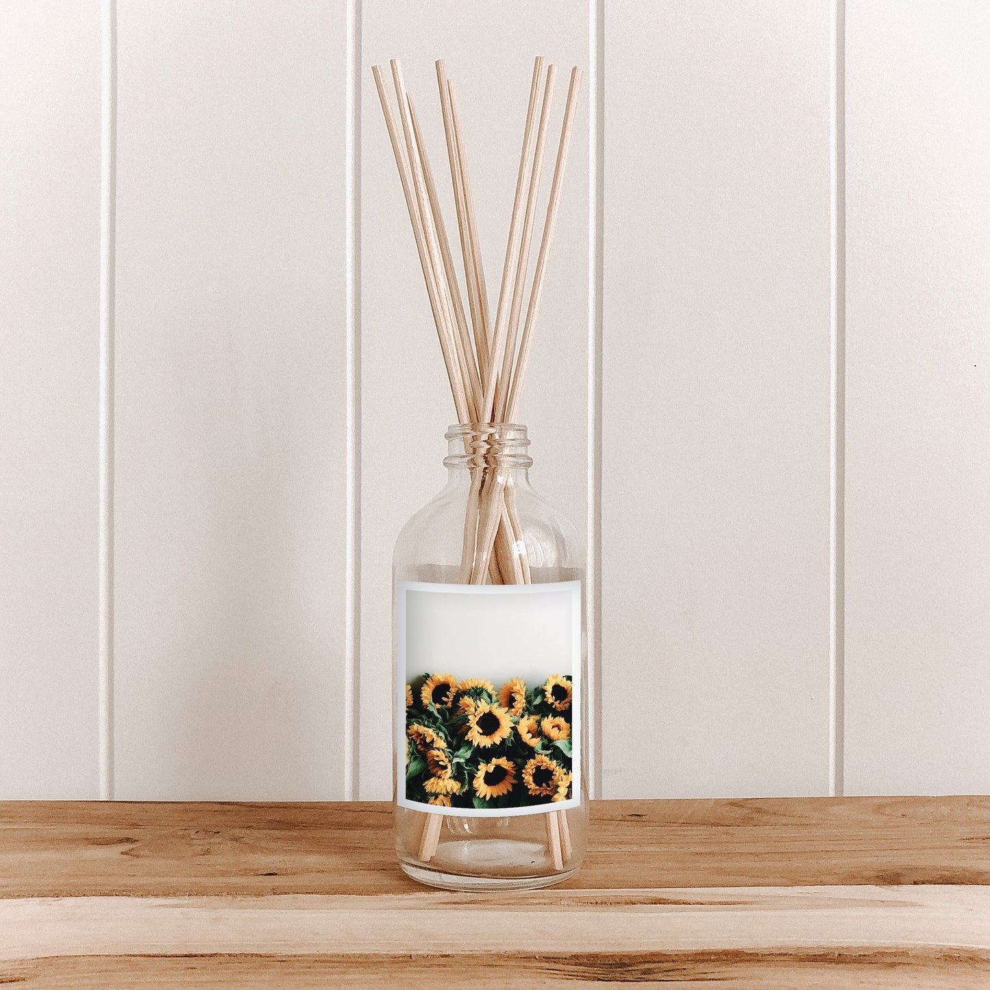 The Commonfolk room diffuser bottle, with a label featuring sunflowers and filled with natural bamboo reed sticks, is placed on a wooden surface against a white background.