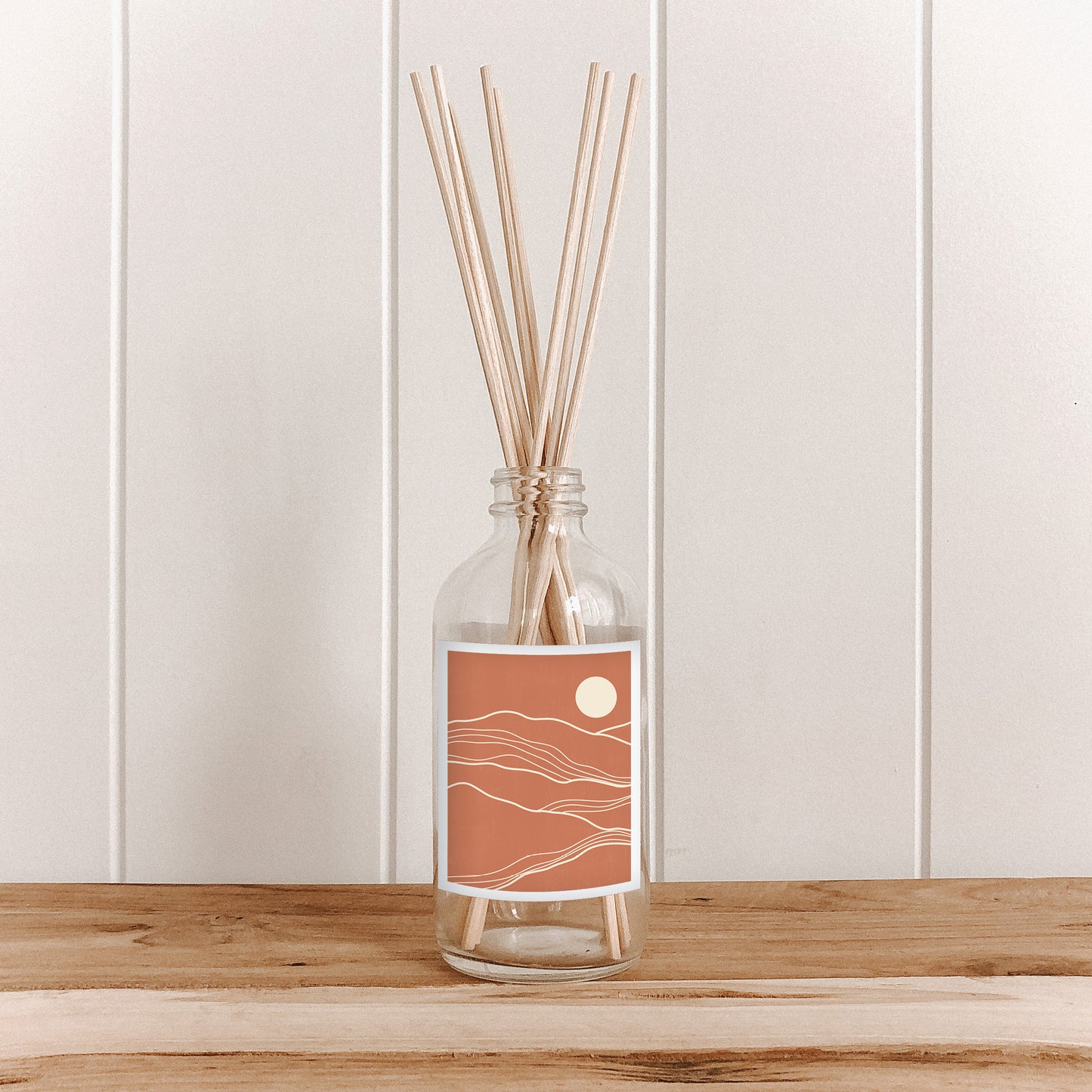 The Commonfolk room diffuser with natural bamboo reed sticks in a clear glass bottle with a terracotta-colored label featuring an abstract landscape.