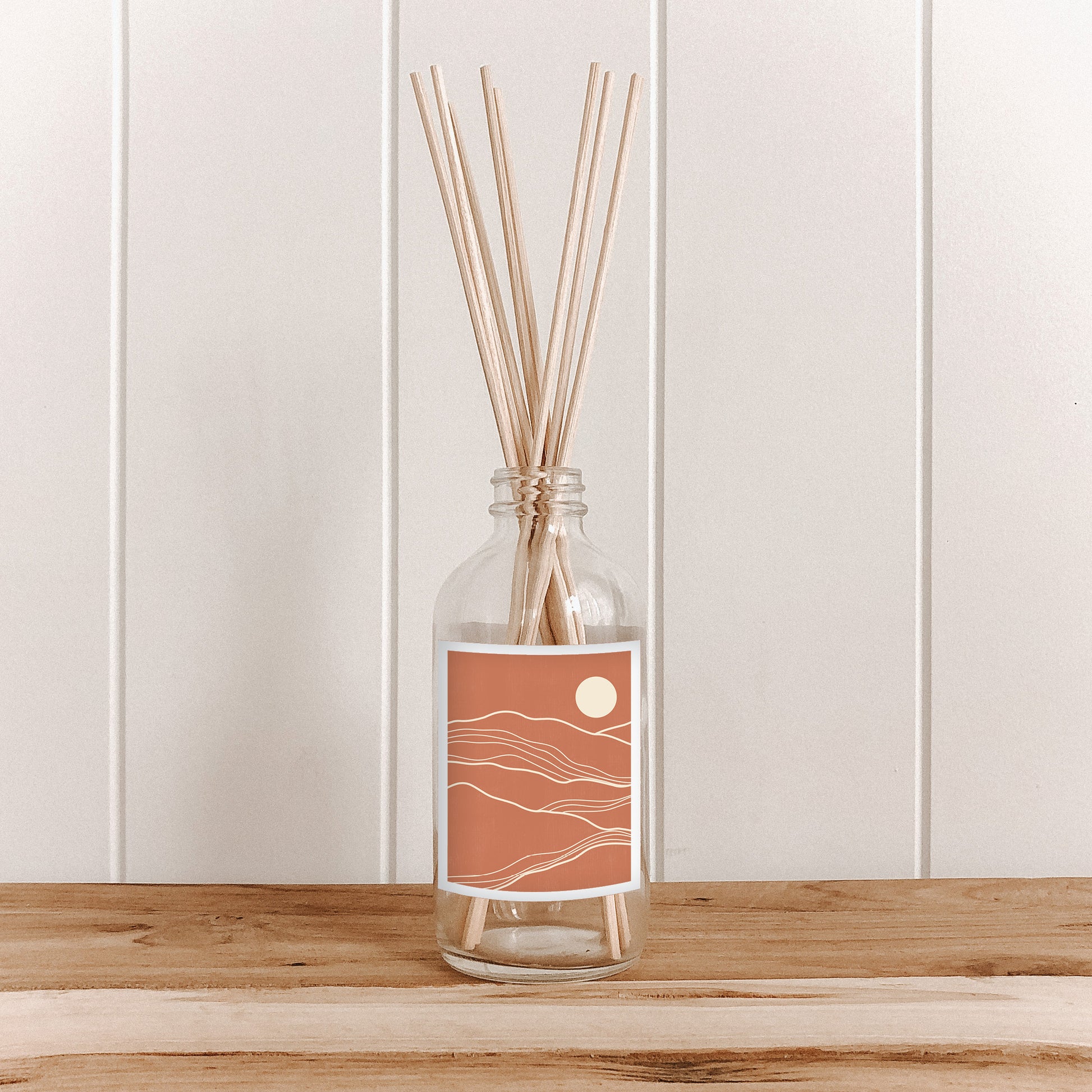 The Commonfolk room diffuser with natural bamboo reed sticks in a clear glass bottle with a terracotta-colored label featuring an abstract landscape.