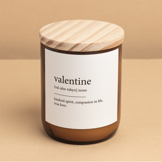 The Commonfolk scented candle has a white label that reads 'valentine' and the definition "kindred spirit, companion in life, true love." The candle is in a brown jar with a wooden lid on top.