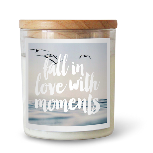 The Commonfolk scented white candle with a wooden lid, a label featuring the text 'Fall in love with moments,' and an image of a beach scene with birds.
