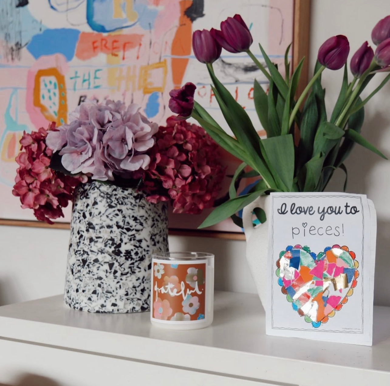 A vase of colorful flowers, alongside a scented candle and a greeting card, is displayed on a wooden shelf.