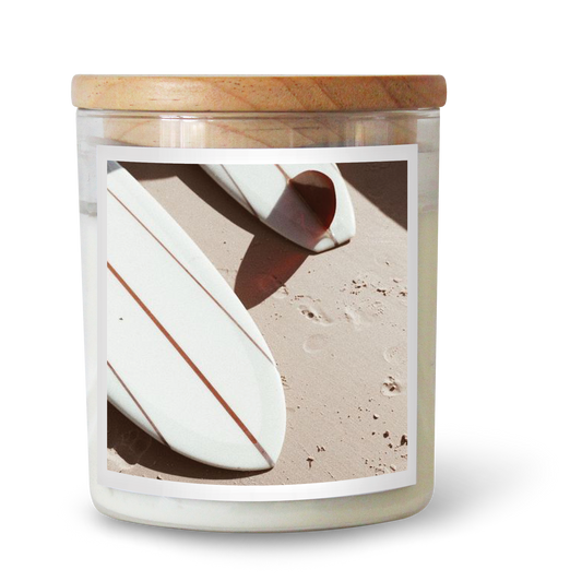 The Commonfolk scented candle features white soy wax, a wooden lid, and a label depicting a white surfboard on a sandy beach.