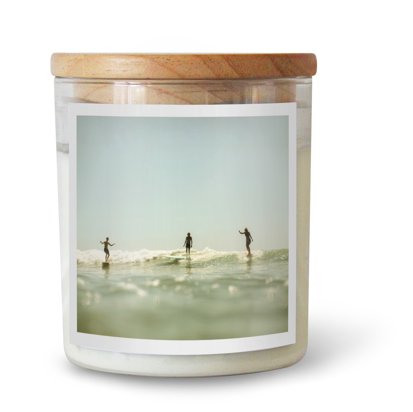 The Commonfolk scented candle has a white wax container featuring an image of three people surfing in the ocean. The candle has a wooden lid and is part of the 'Party Wave' collection.