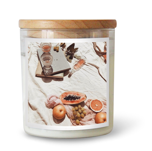 The Commonfolk scented candle has a label featuring a picnic scene, including fruits and a glass of wine, with a wooden lid on top.