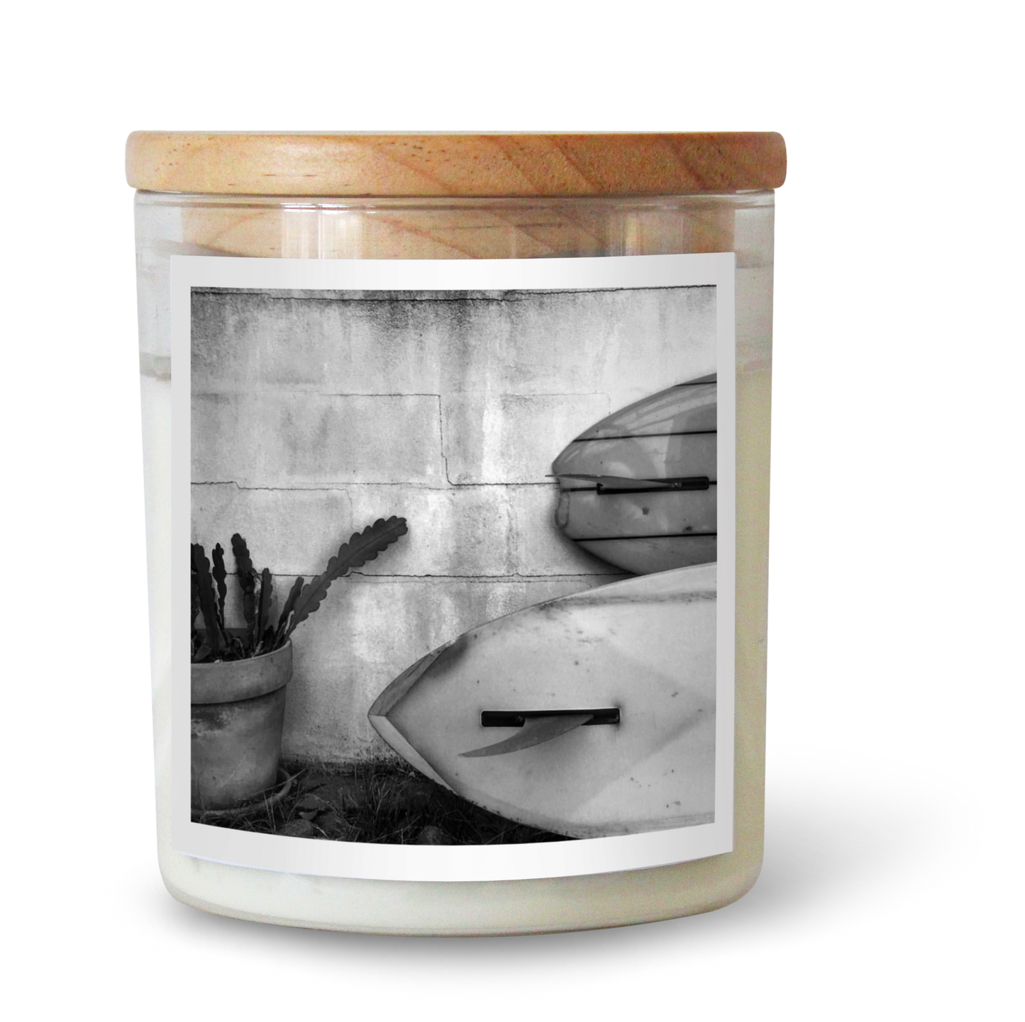 The Commonfolk scented candle comes in a clear jar featuring a black and white photo of a surfboard and beach scene on the label, topped with a wooden lid.
