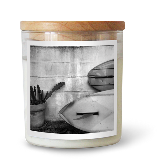 The Commonfolk scented candle comes in a clear jar featuring a black and white photo of a surfboard and beach scene on the label, topped with a wooden lid.