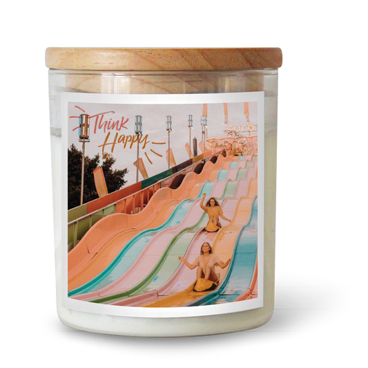 The Commonfolk scented white candle has a label featuring an illustration of a slide and the text 'Think Happy.' The candle has a wooden lid.