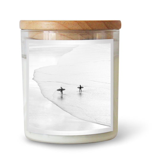 The Commonfolk scented candle has white wax, housed in a clear glass jar with a wooden lid. The wick appears to be in a surf shape, and there are decorative surfboard-like figures beside the jar.