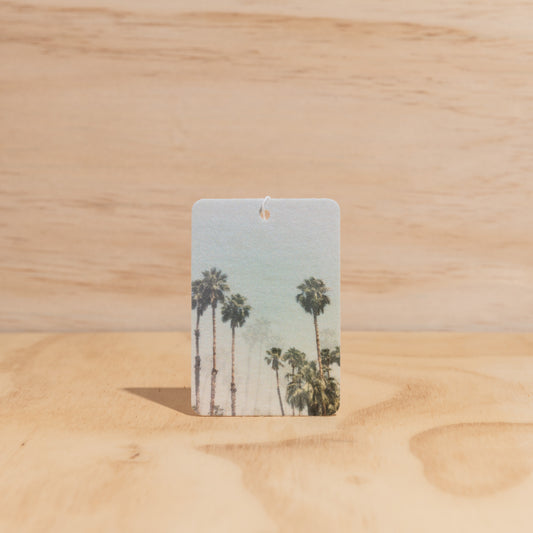 The Commonfolk air freshener, with a printed image of palm trees against a blue sky, is displayed on a wooden surface.