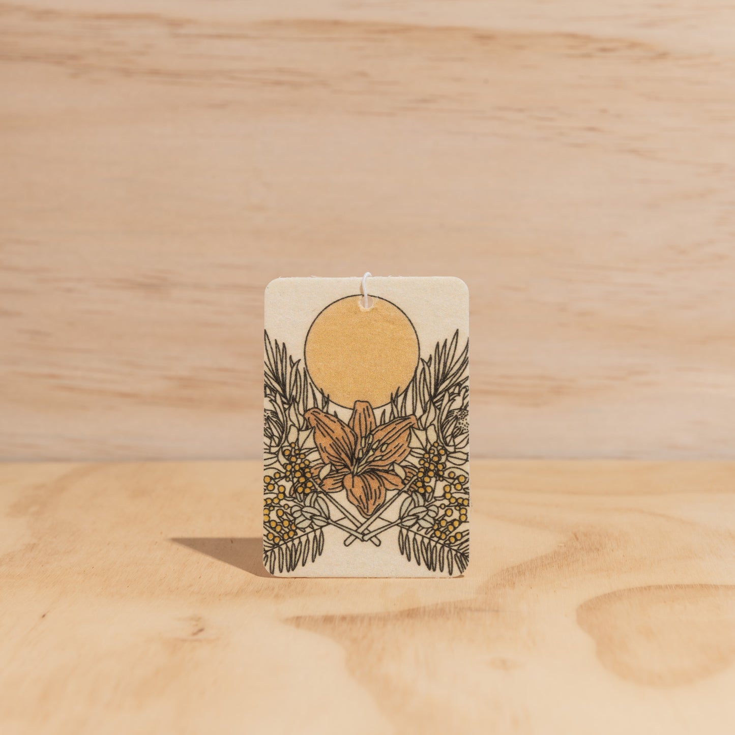 The Commonfolk air freshener, with a floral design predominantly in gold and white colors, is set against a wooden background.