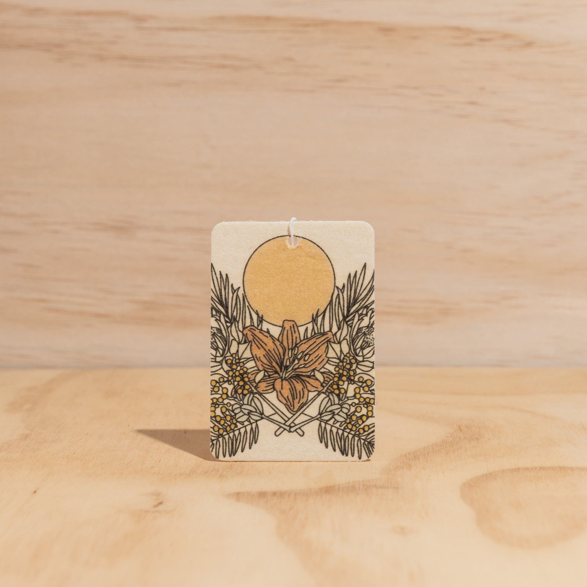 The Commonfolk air freshener, with a floral design predominantly in gold and white colors, is set against a wooden background.