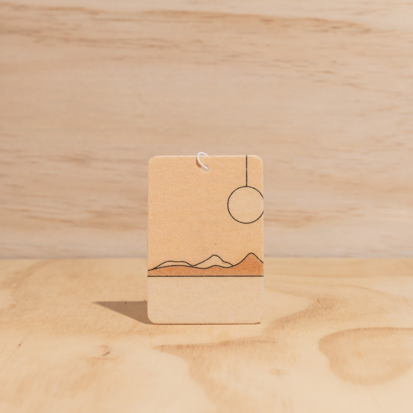 The Commonfolk air freshener, with a simple design featuring sun and mountain-like shapes in a brown color, displays against a wooden background.