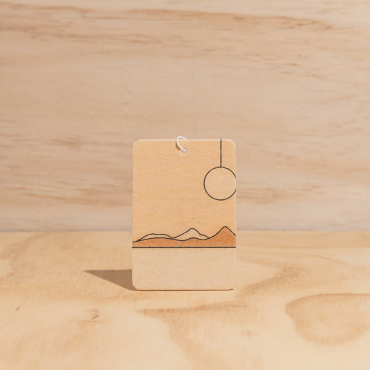 The Commonfolk air freshener, with a simple design featuring sun and mountain-like shapes in a brown color, displays against a wooden background.