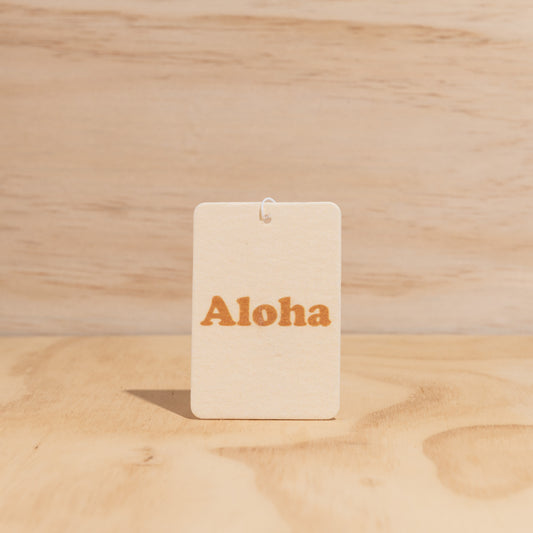 The Commonfolk air freshener with the word 'Aloha' printed in orange displays against a wooden background.