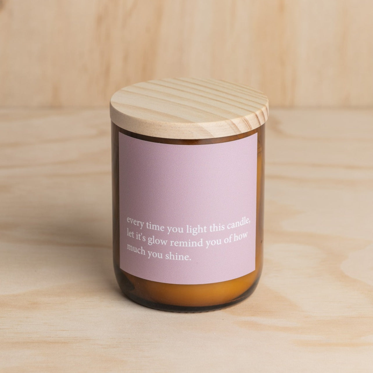 The Commonfolk scented soy candle in an amber jar with a wooden lid, features a purple label with white text that reads 'every time you light this candle, let its glow remind you of how much you shine.'.